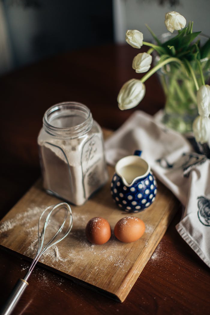 From above of vase with white flowers placed near fresh milk eggs and glass jar with flour on cutting woody board at kitchen