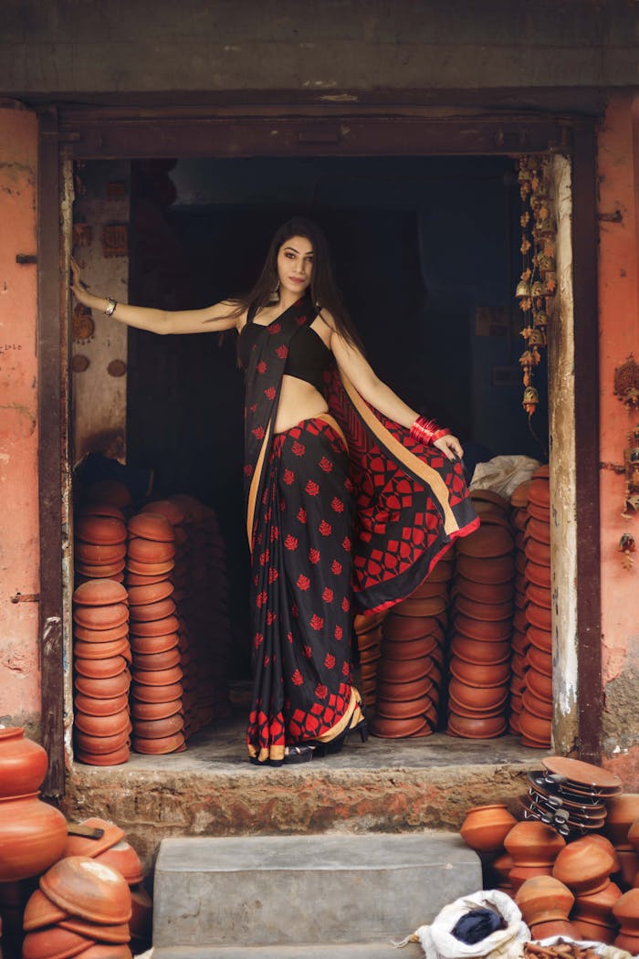 A woman in a black saree poses gracefully amidst stacks of pottery, evoking traditional elegance.
